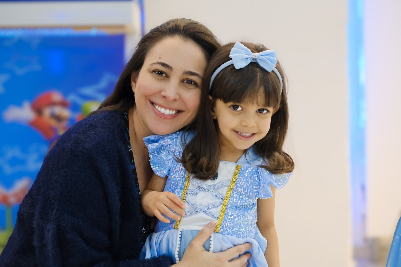 Mãe abraçando a filha vestida de Cinderela, as duas sorrindo, numa festa infantil