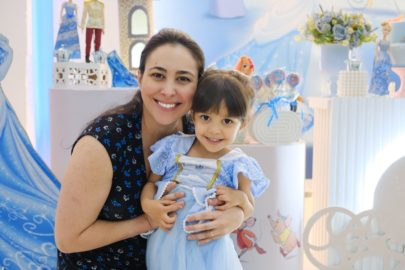 Mãe agachada abraçando a filha de vestido azul, as duas sorrindo para a câmera