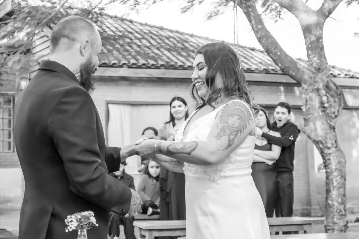 Noivos de mãos dadas trocando as alianças diante dos convidados, em preto e branco