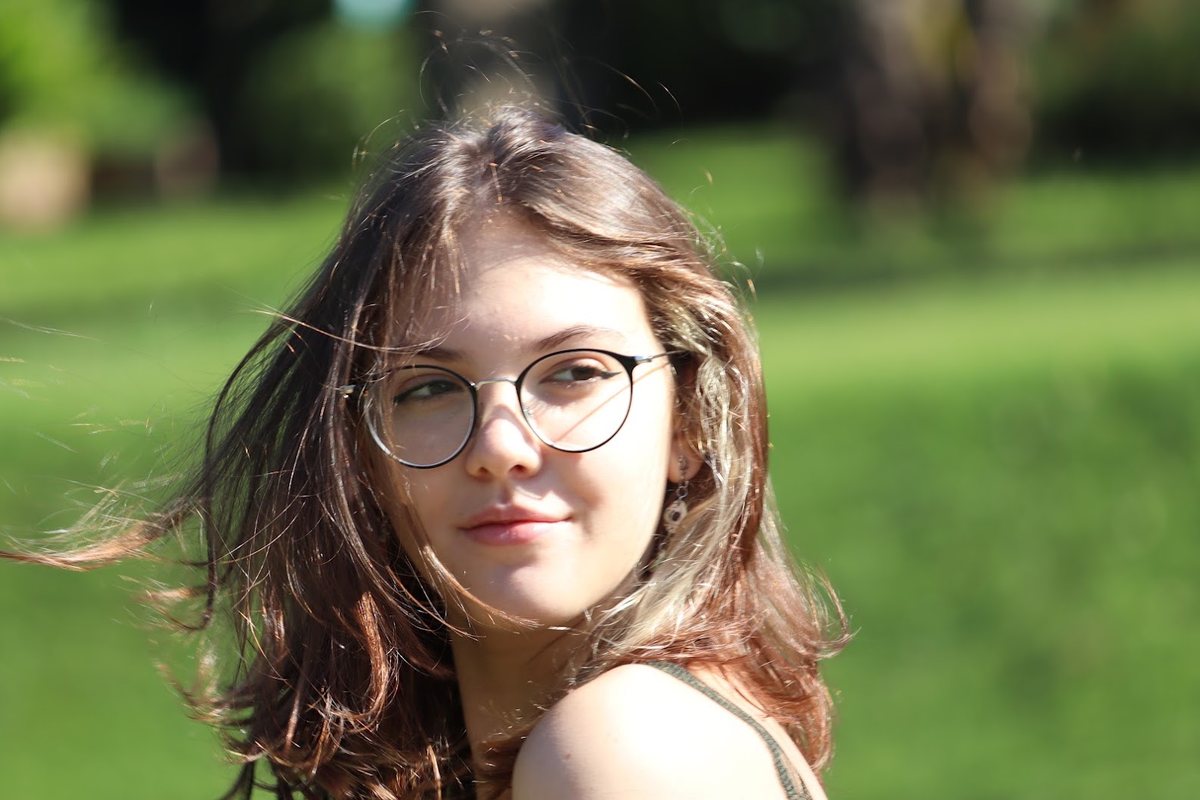 Moça de óculos e cabelo ao vento sorrindo, num gramado com luz de fim de tarde