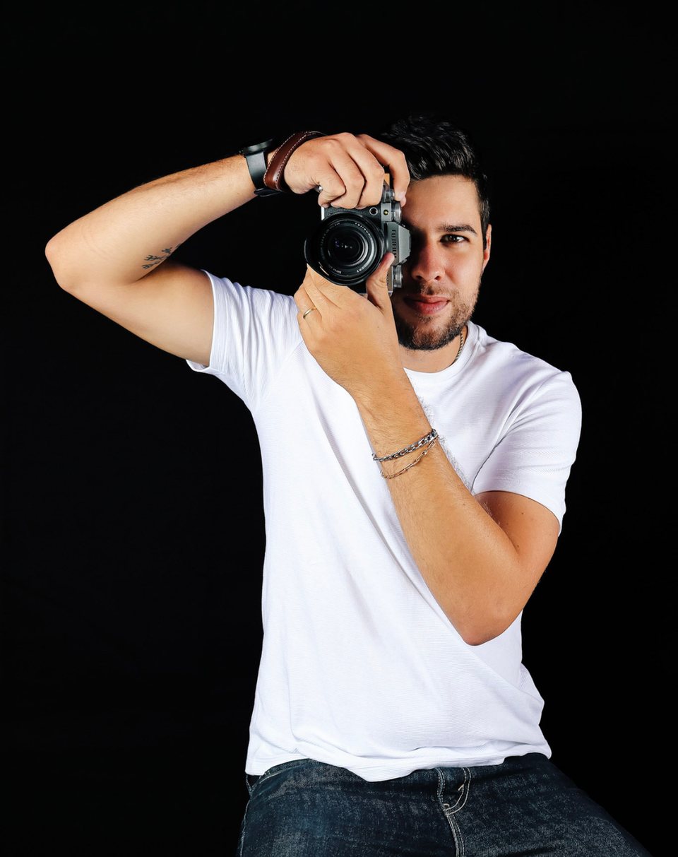 Hugo, fotógrafo da LHG, de camiseta branca, mirando a câmera com um olho só, em fundo preto