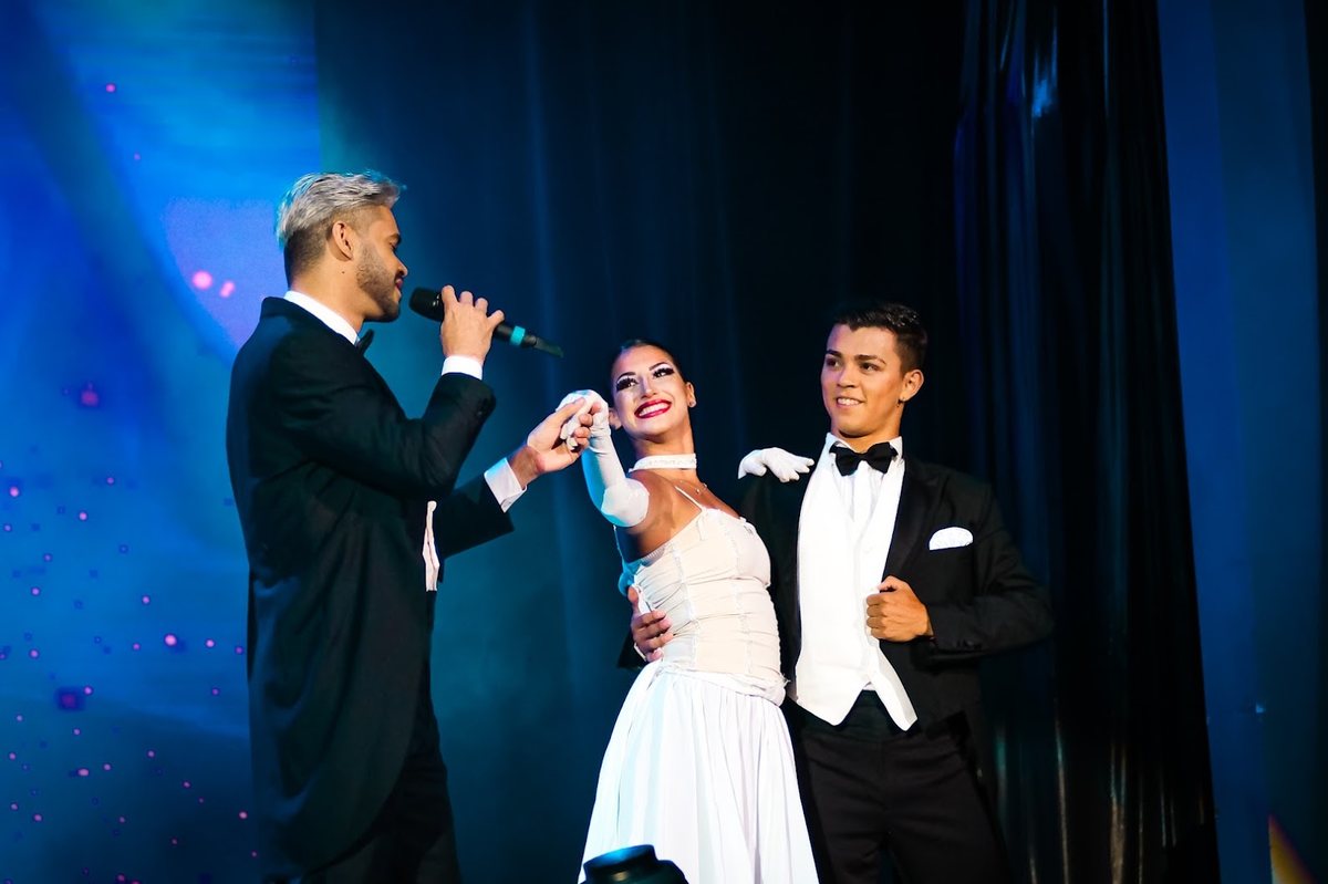 Cantor de fraque cantando para uma bailarina de branco no palco, com outro bailarino ao lado